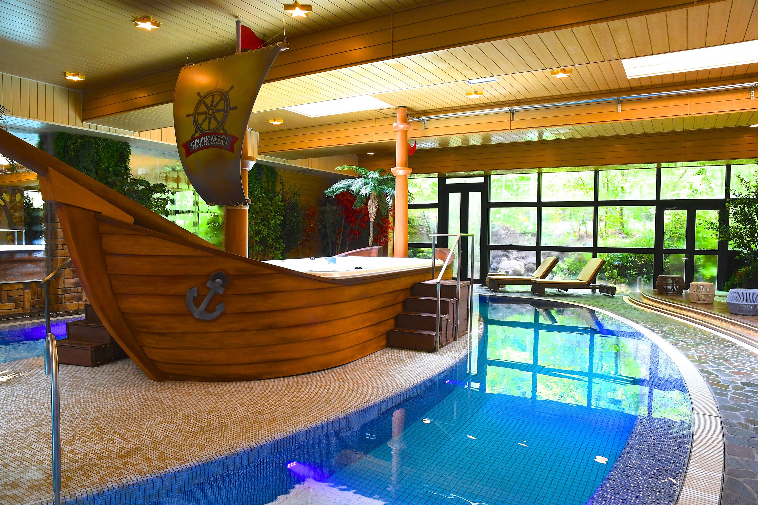 A pool area with a ship-shaped deck, a hot tub, and a view of greenery outside.