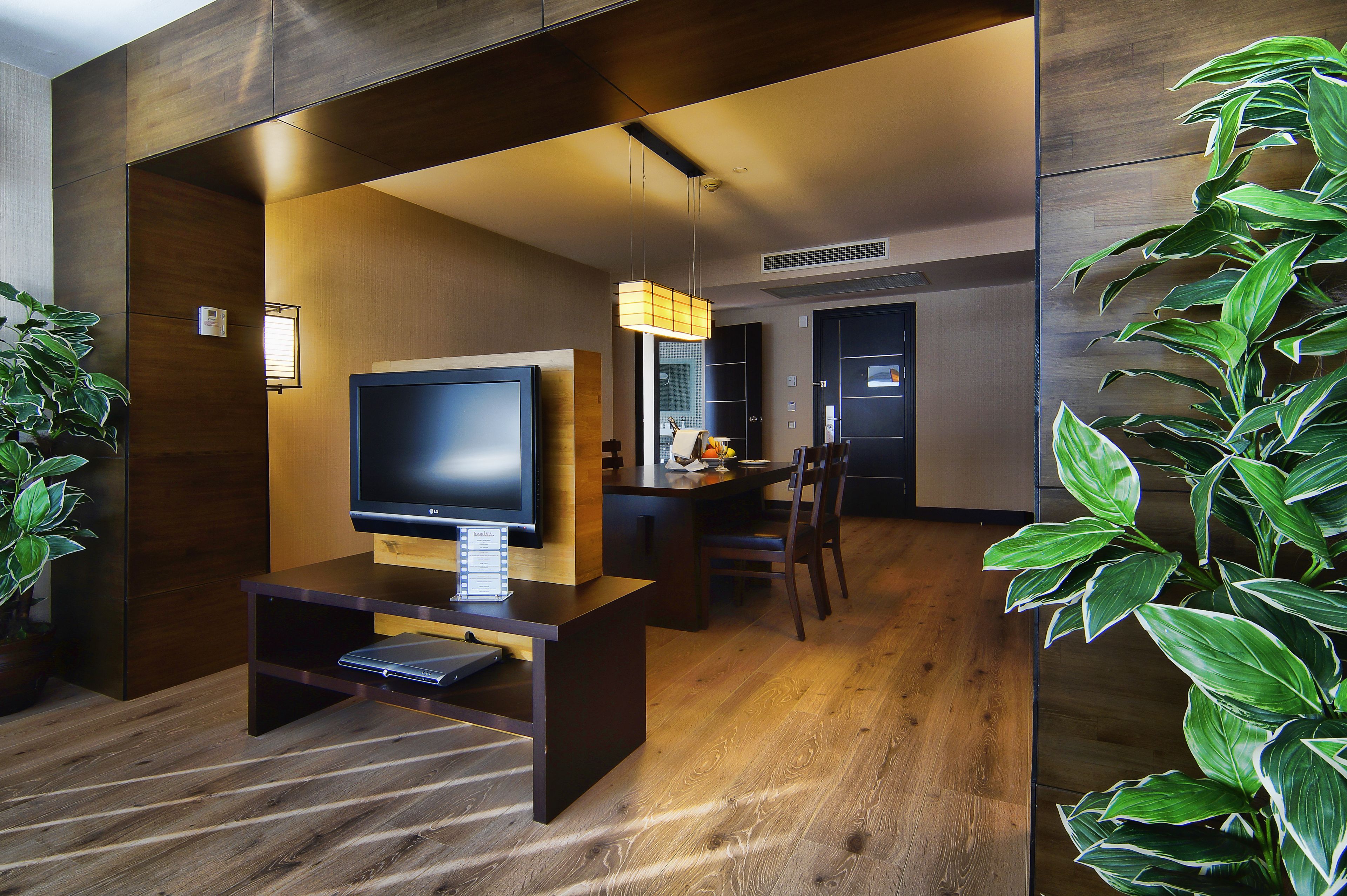 Een moderne hotelkamer met een houten vloer, een flatscreen televisie op een houten standaard, een zithoek met een tafel en stoelen, en een grote potplant.