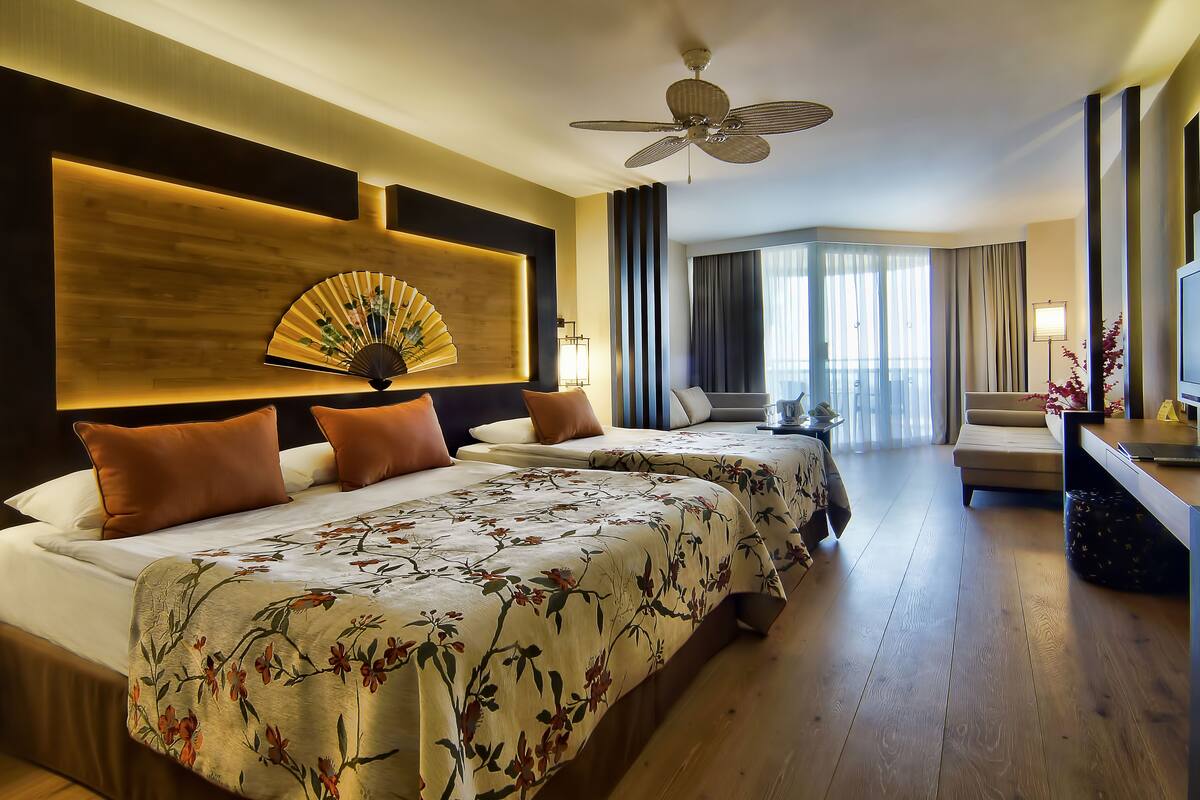 A hotel room with two beds, a ceiling fan, a large window with curtains, and a flat-screen TV.