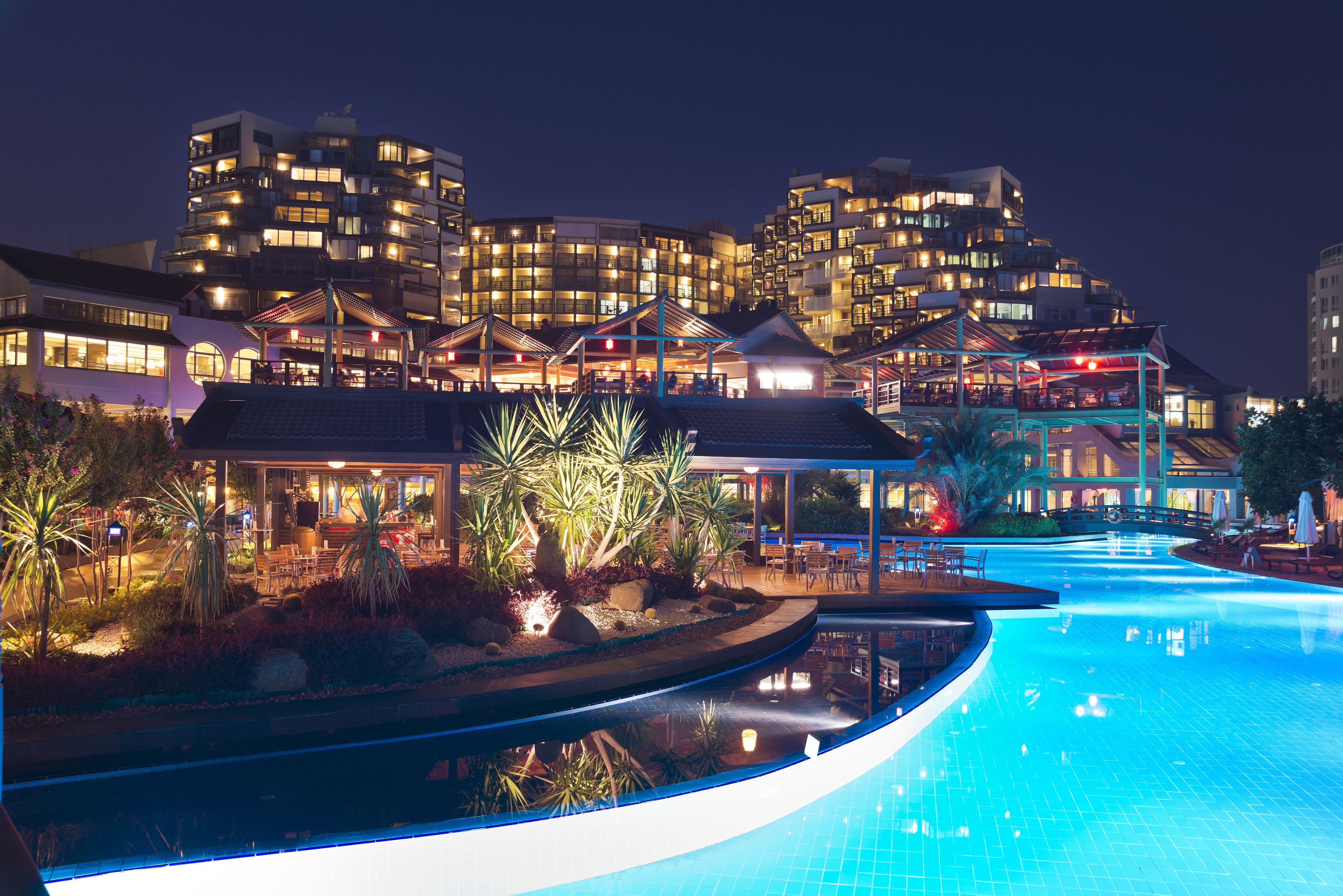 Vista nocturna de un hotel con piscina, zonas de estar al aire libre y un diseño arquitectónico moderno.