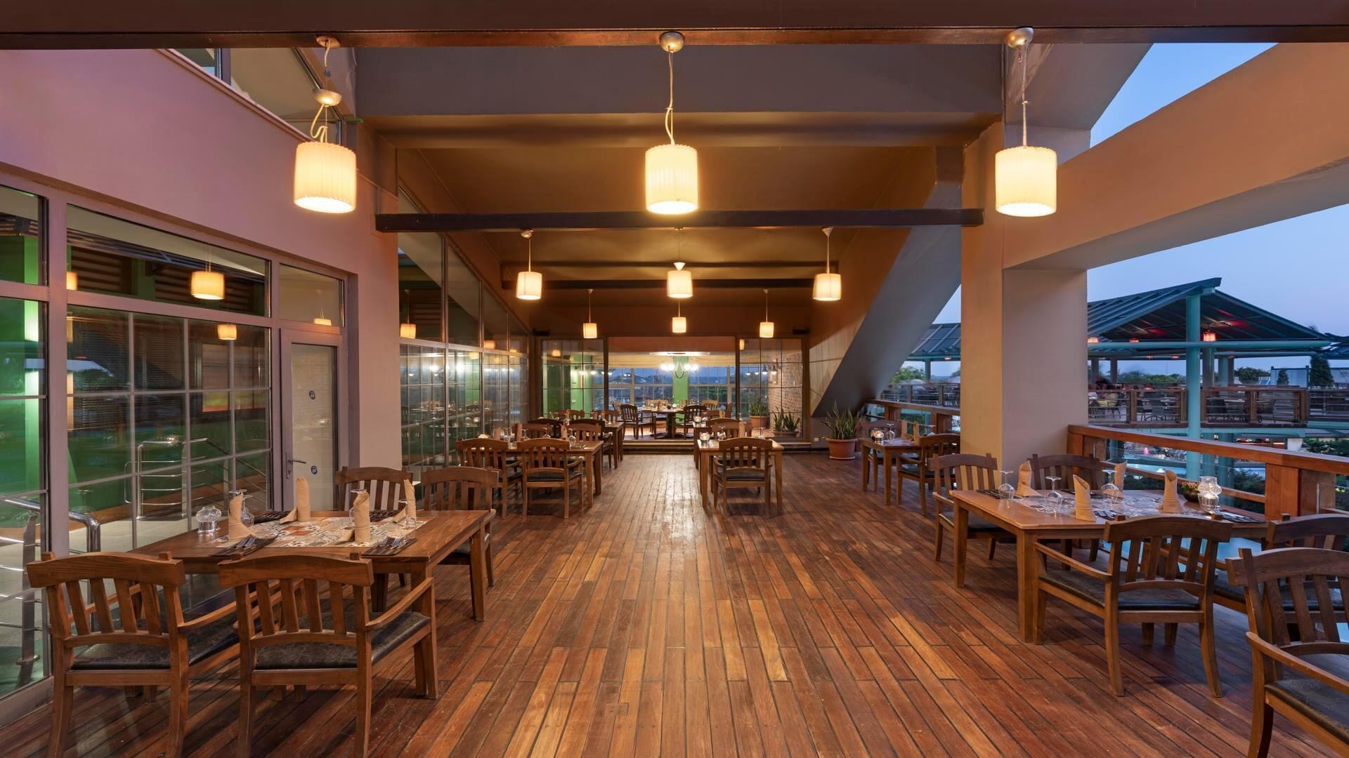 An indoor dining area with wooden tables and chairs, large windows, and hanging pendant lights.