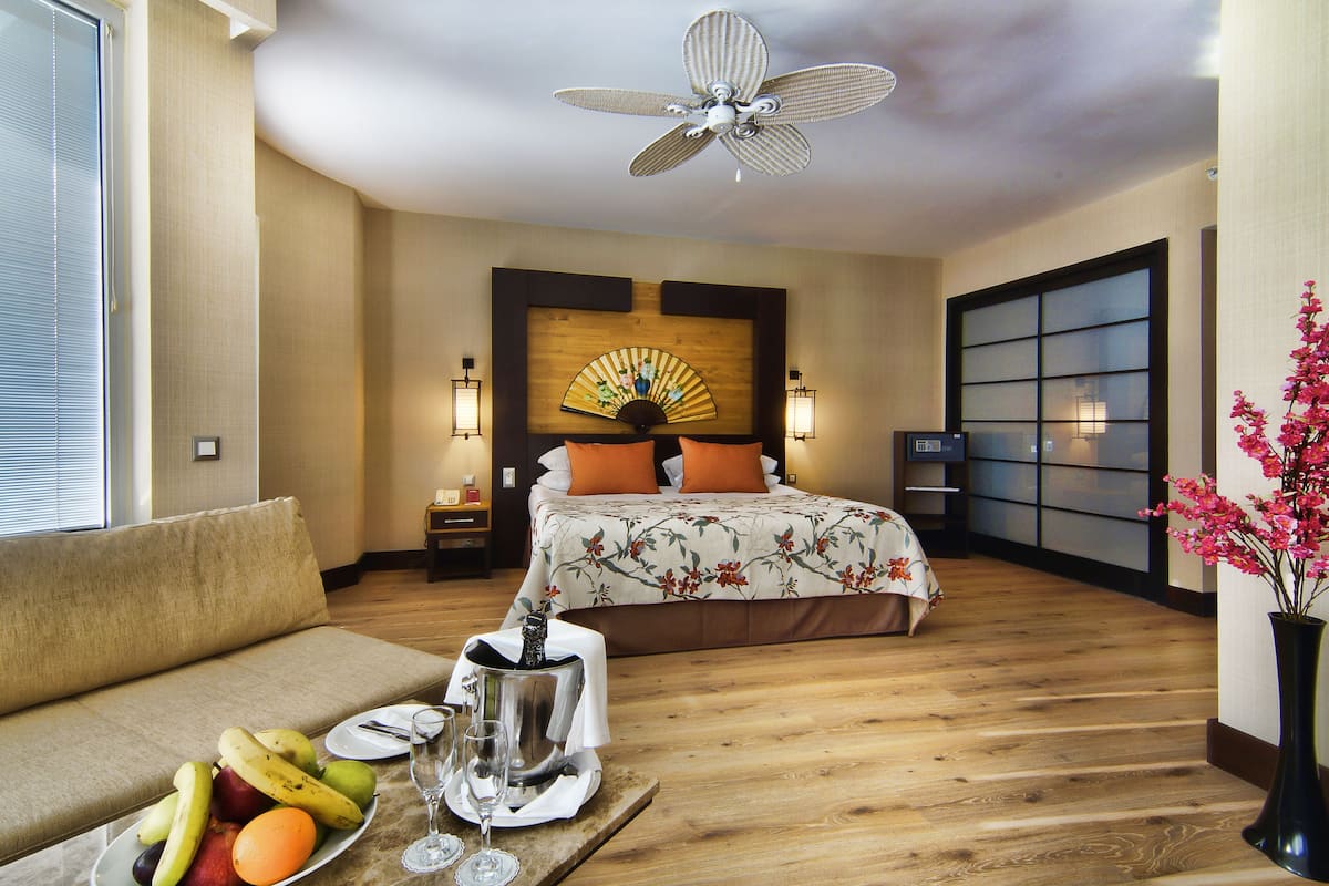 A hotel room with a bed, a bedside table, a nightstand, a fan, a window, a sofa, a tray with fruit and glasses, and a vase with flowers.