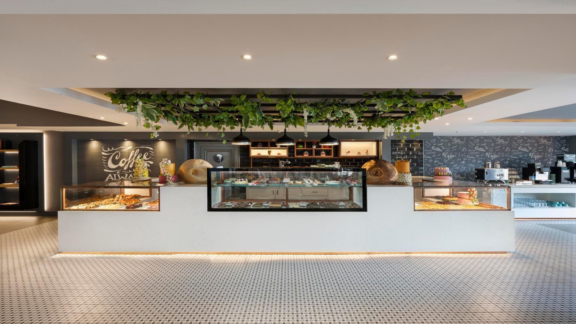Una cafetería moderna con un mostrador central que exhibe pasteles y postres, rodeada de estanterías y una pared de pizarra con palabras relacionadas con el café.