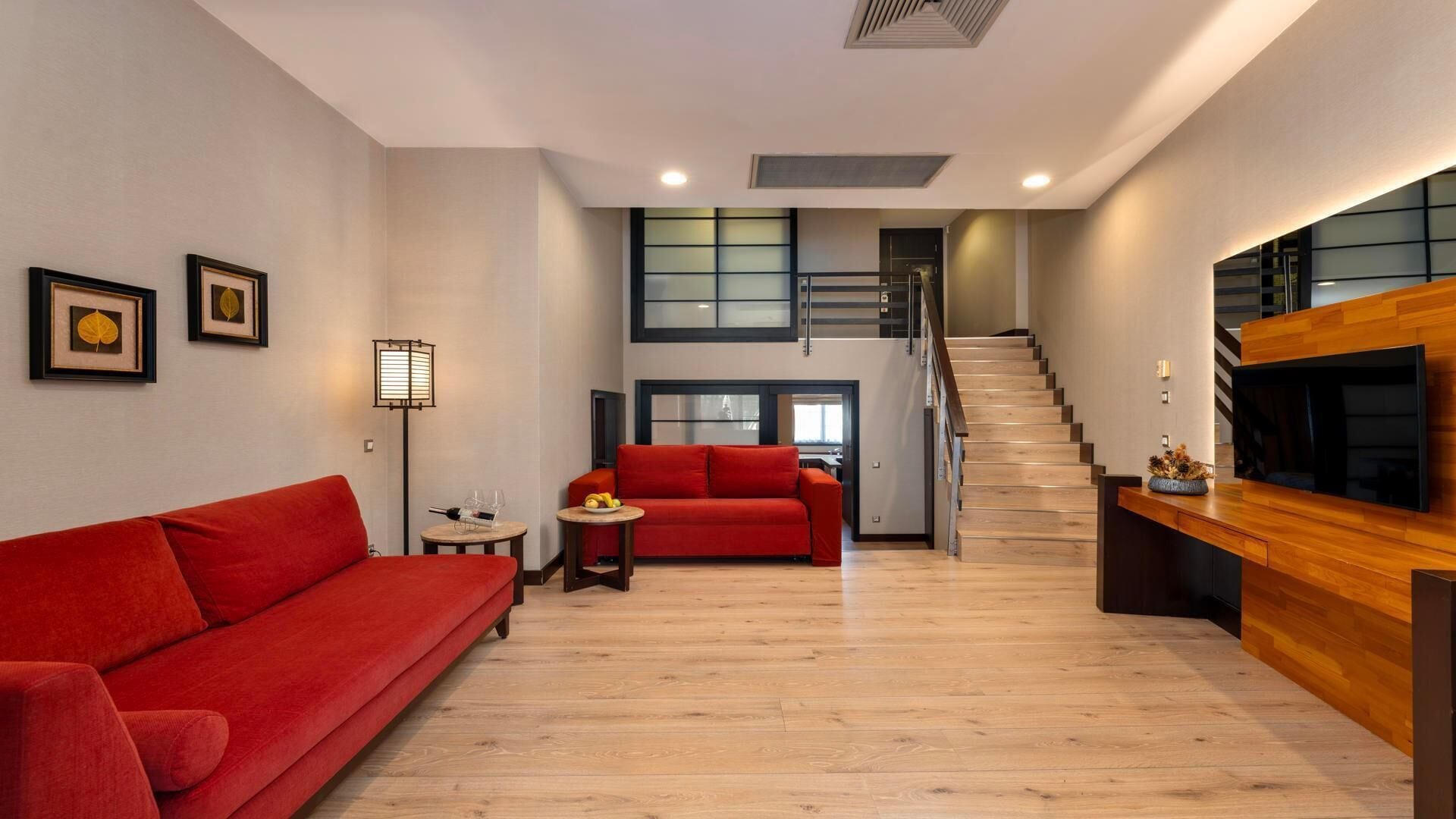 A modern living room with a red sofa, a small round table, and a TV mounted on a wooden wall. A staircase leads to an upper level.