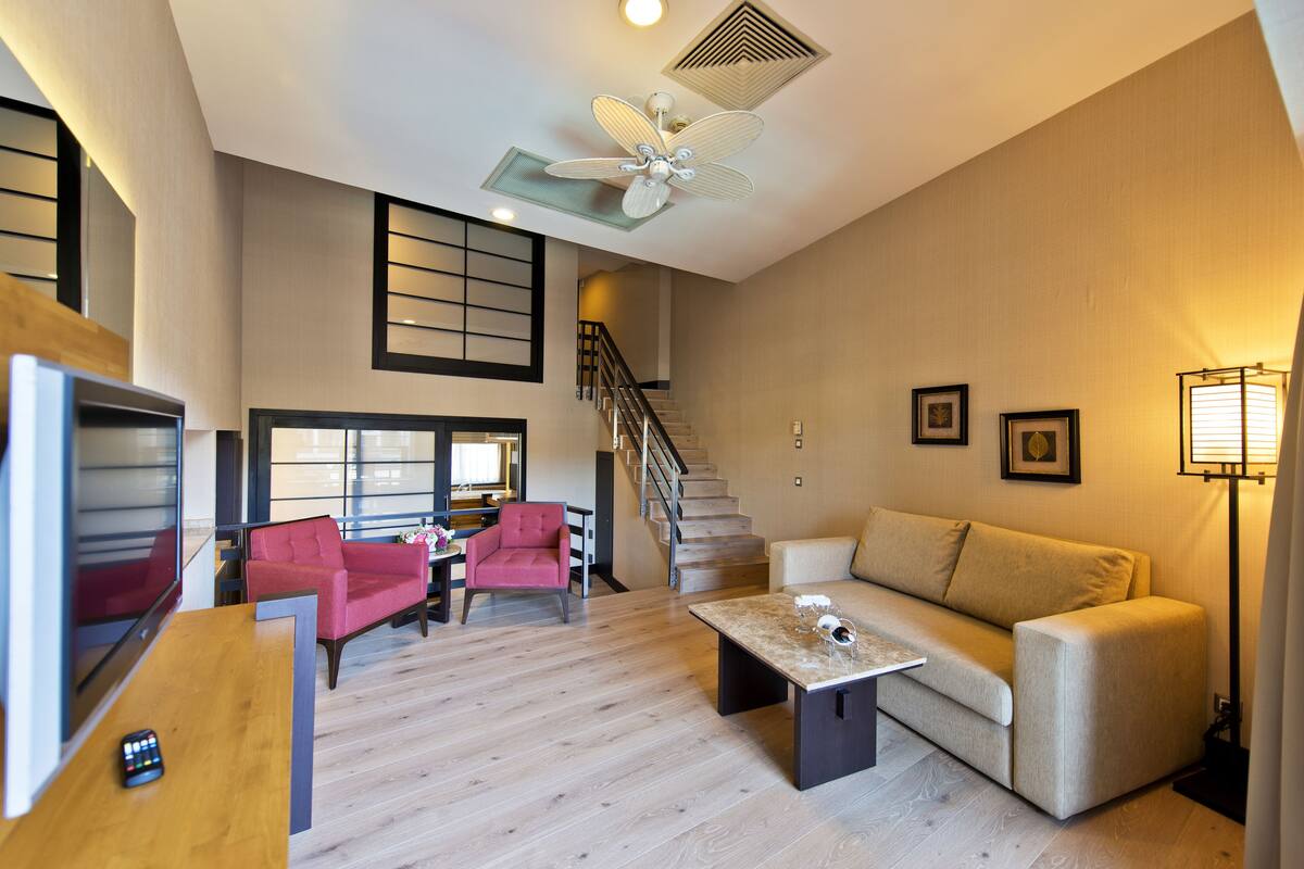 A spacious living room with a wooden floor, a beige sofa, a red armchair, and a coffee table. A ceiling fan and a staircase are visible in the background.