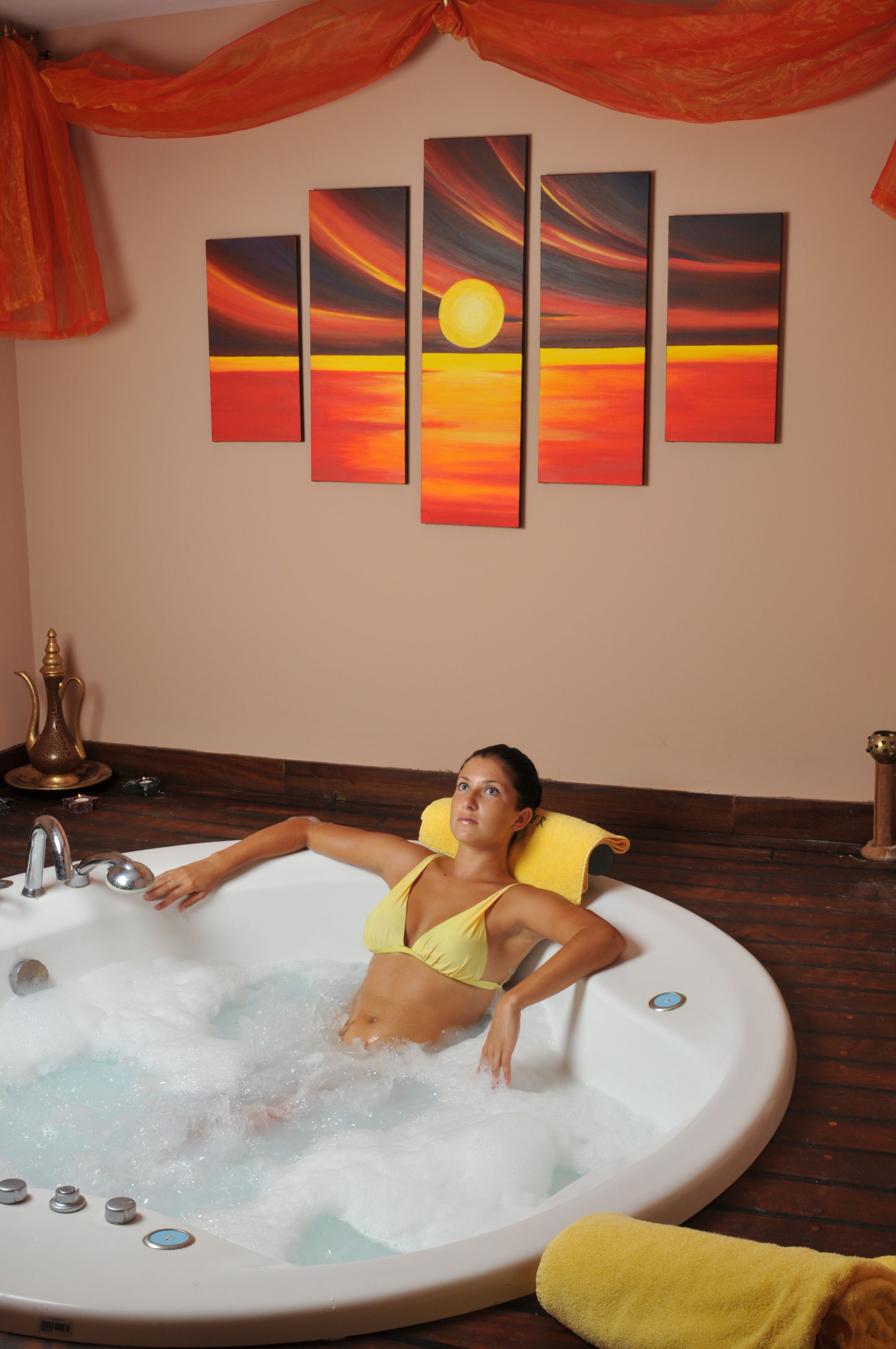 A woman in a yellow swimsuit relaxing in a bathtub with a sunset-themed wall art above.