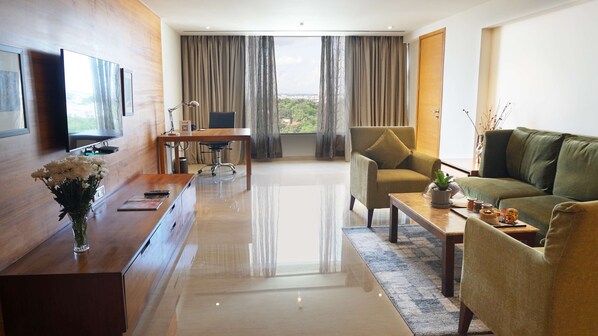 A modern hotel room with a wooden desk, a sofa, a TV mounted on the wall, and a large window with curtains.