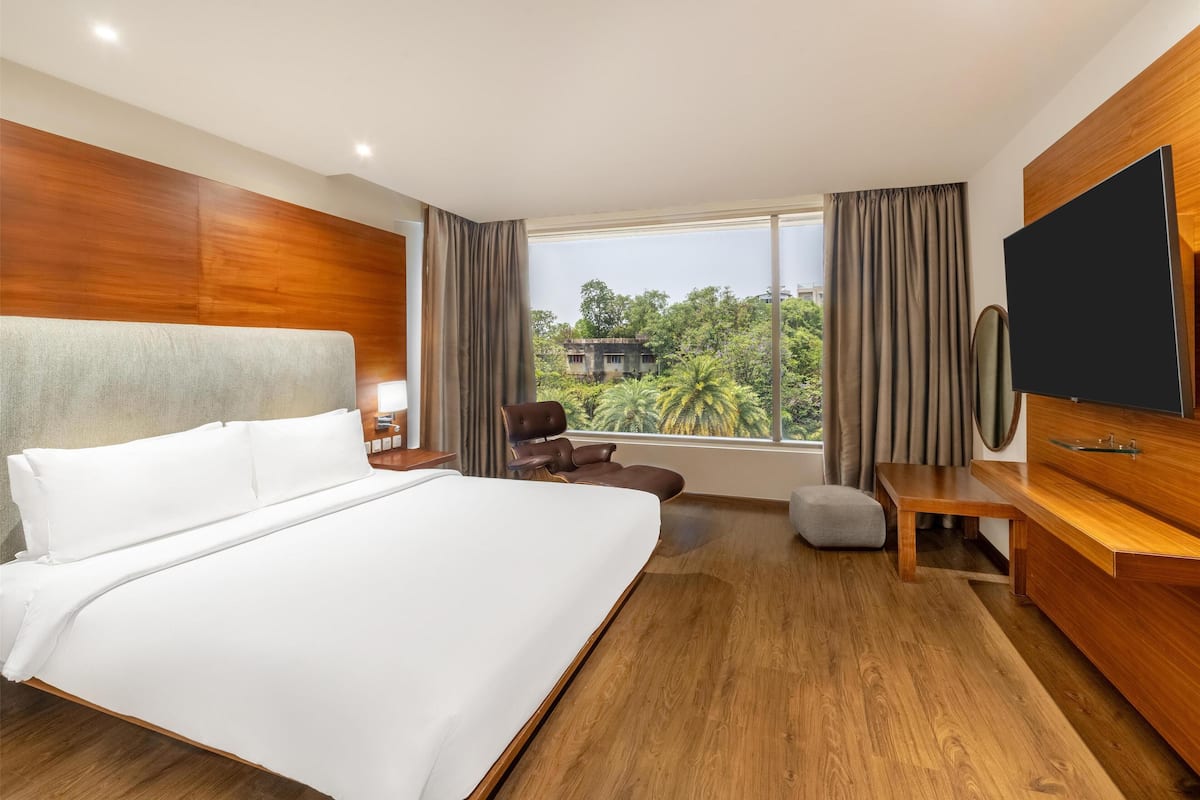 Ein Hotelzimmer mit einem großen Bett, einem Holzkopfstück, einem Fernseher, einem Schreibtisch und Blick auf Grün.