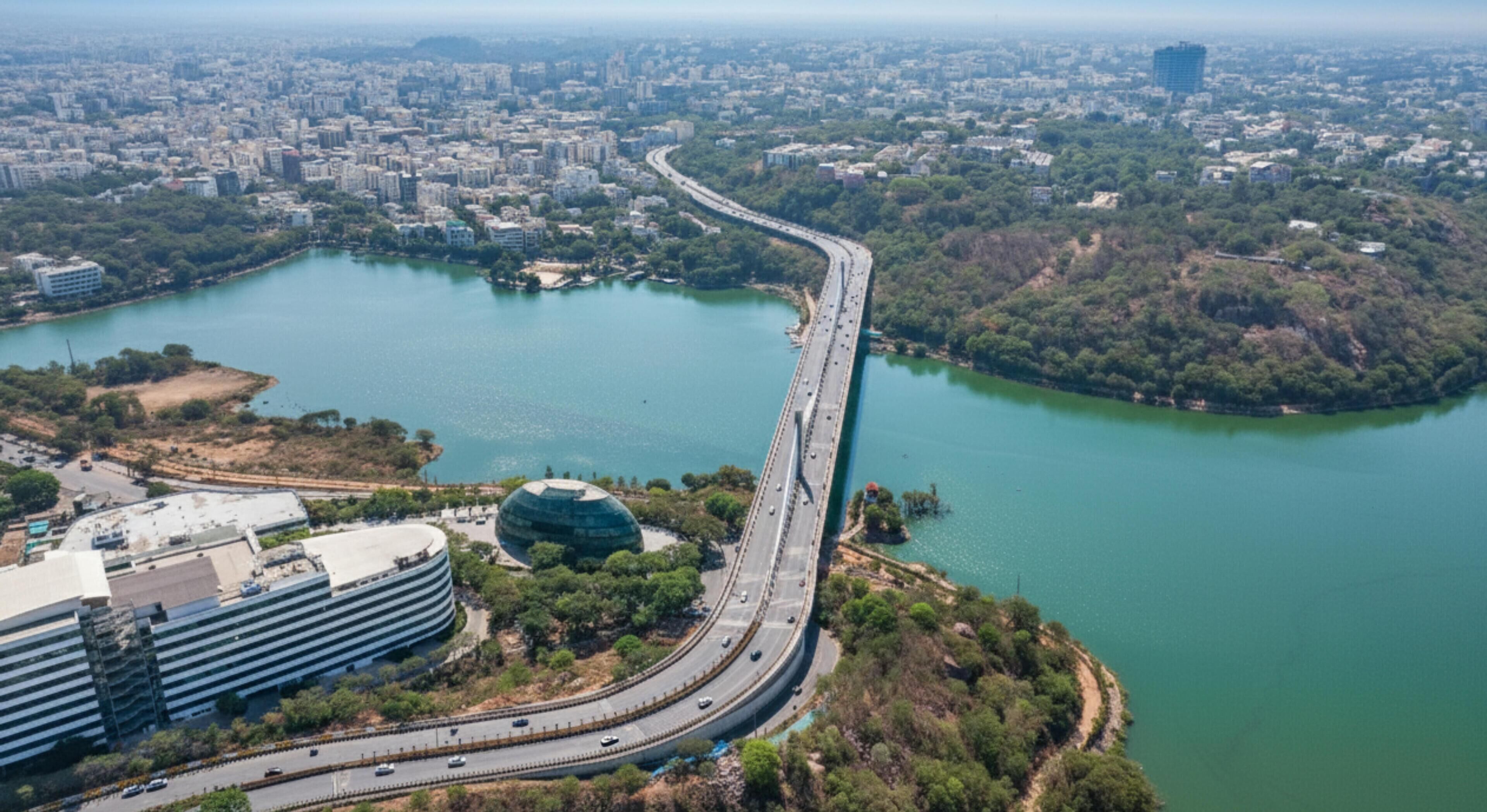 Une vue aérienne d’une ville, avec un pont imposant qui enjambe un vaste plan d’eau, le tout étant entouré de quartiers urbains denses et d’espaces verts.