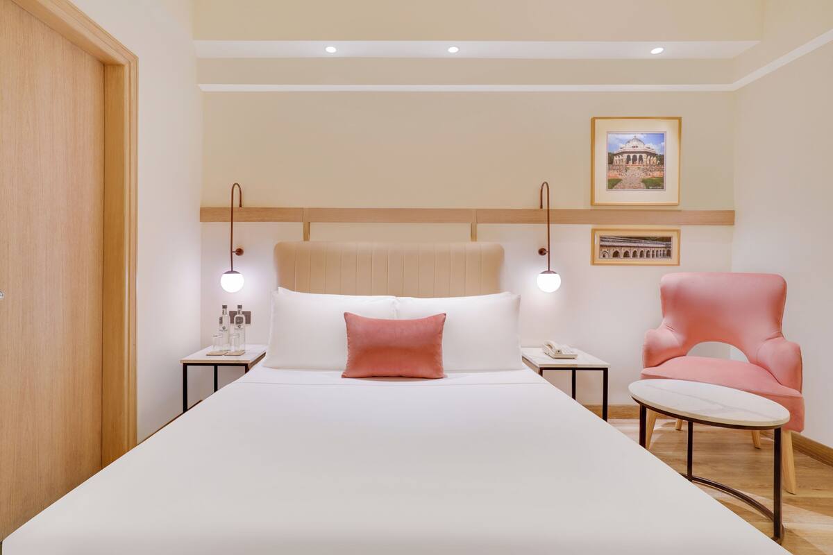 Ein Hotelzimmer mit einem großen Bett, einem hölzernen Kopfteil, Nachttischen mit Lampen, einem rosa Sessel und gerahmten Bildern an der Wand.