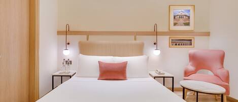 A hotel room with a large bed, a wooden headboard, bedside tables with lamps, a pink armchair, and framed pictures on the wall.