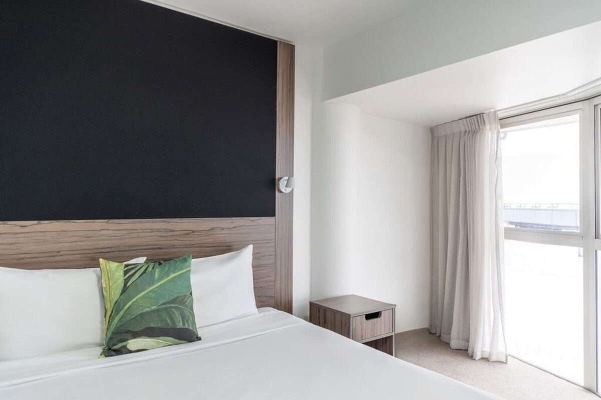 A hotel room with a bed, a nightstand, a window with curtains, and a view of the outside.