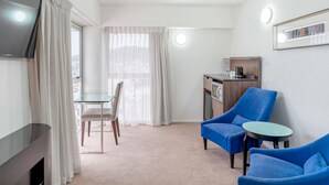 A hotel room with a TV, a dining table with chairs, a kitchenette, and two blue armchairs.