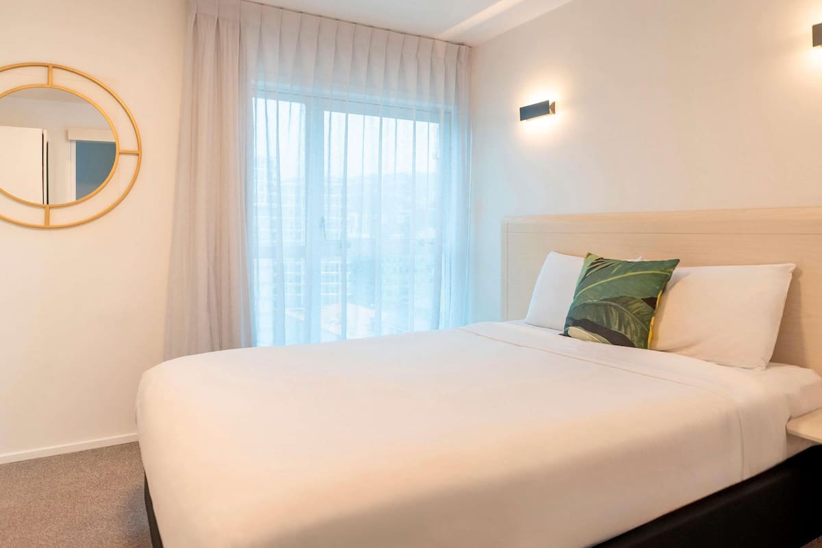 Ein Hotelzimmer mit Bett, rundem Spiegel, Fenster mit Vorhängen und Wandleuchte.