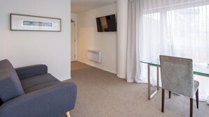 A modern hotel room with a grey sofa, a glass dining table, a TV mounted on the wall, and a large window with sheer curtains.