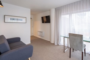 Camera d'hotel moderna con un divano grigio, un tavolo da pranzo in vetro, una TV a parete e un'ampia finestra con tende leggere.