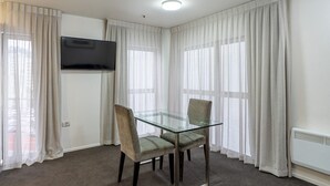 A room with a glass-top table, two chairs, a flat-screen TV mounted on the wall, and large windows with sheer curtains.