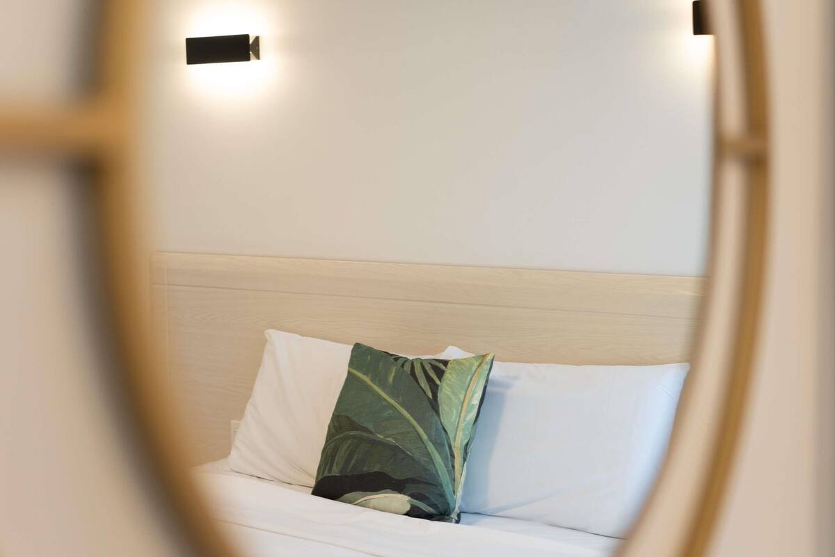 A bed with white bedding and a green leaf-patterned pillow, a wooden headboard, and two wall-mounted light fixtures.