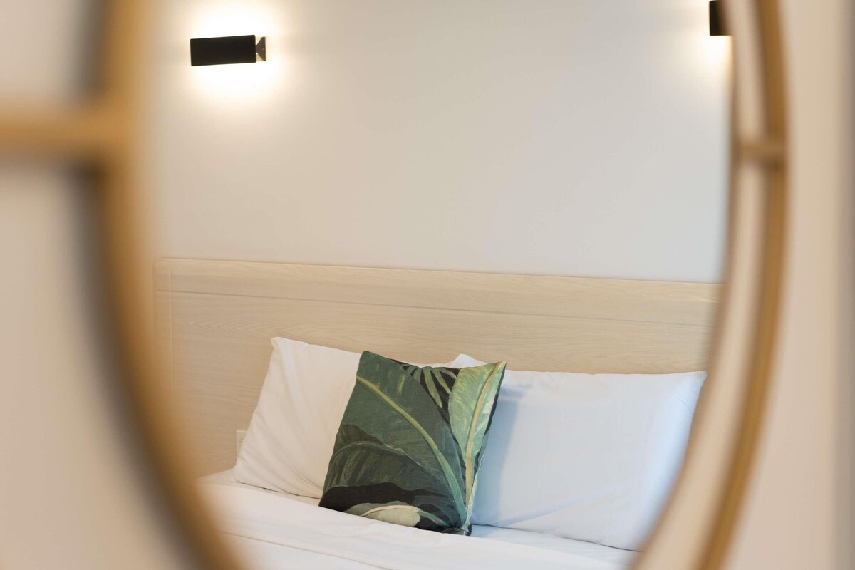 A bed with white bedding and a green leaf-patterned pillow, a wooden headboard, and two wall-mounted light fixtures.