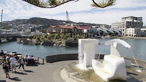 A waterfront park with a modern fountain, a bridge, and buildings in the background.