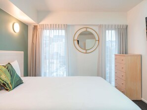 Hotellihuone, jossa on sänky, lipasto, pyöreä peili ja ikkuna, jossa on verhot.
