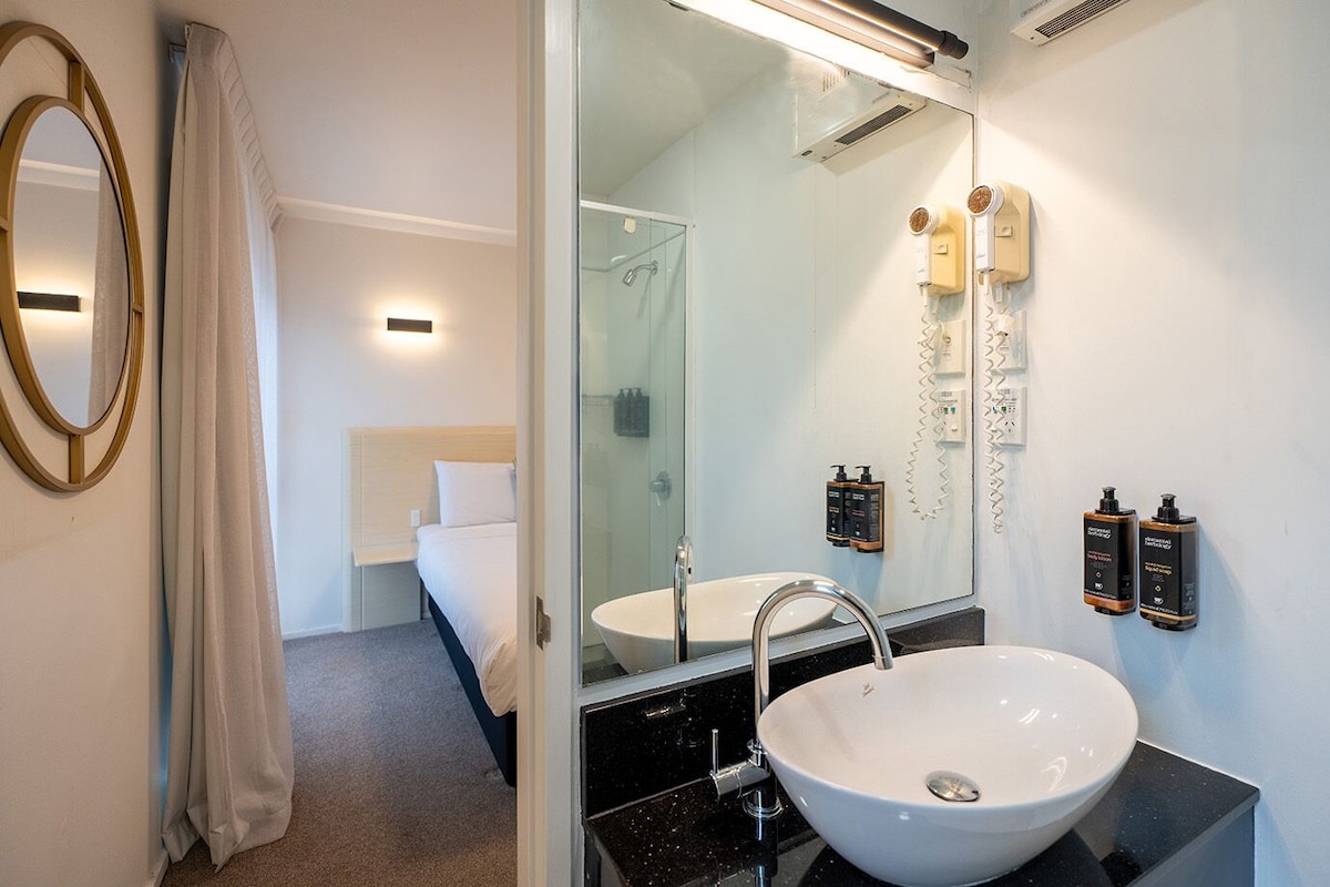 A modern bathroom with a large mirror, a white sink, and a shower. There are two wall-mounted hair dryers and two bottles of shampoo.