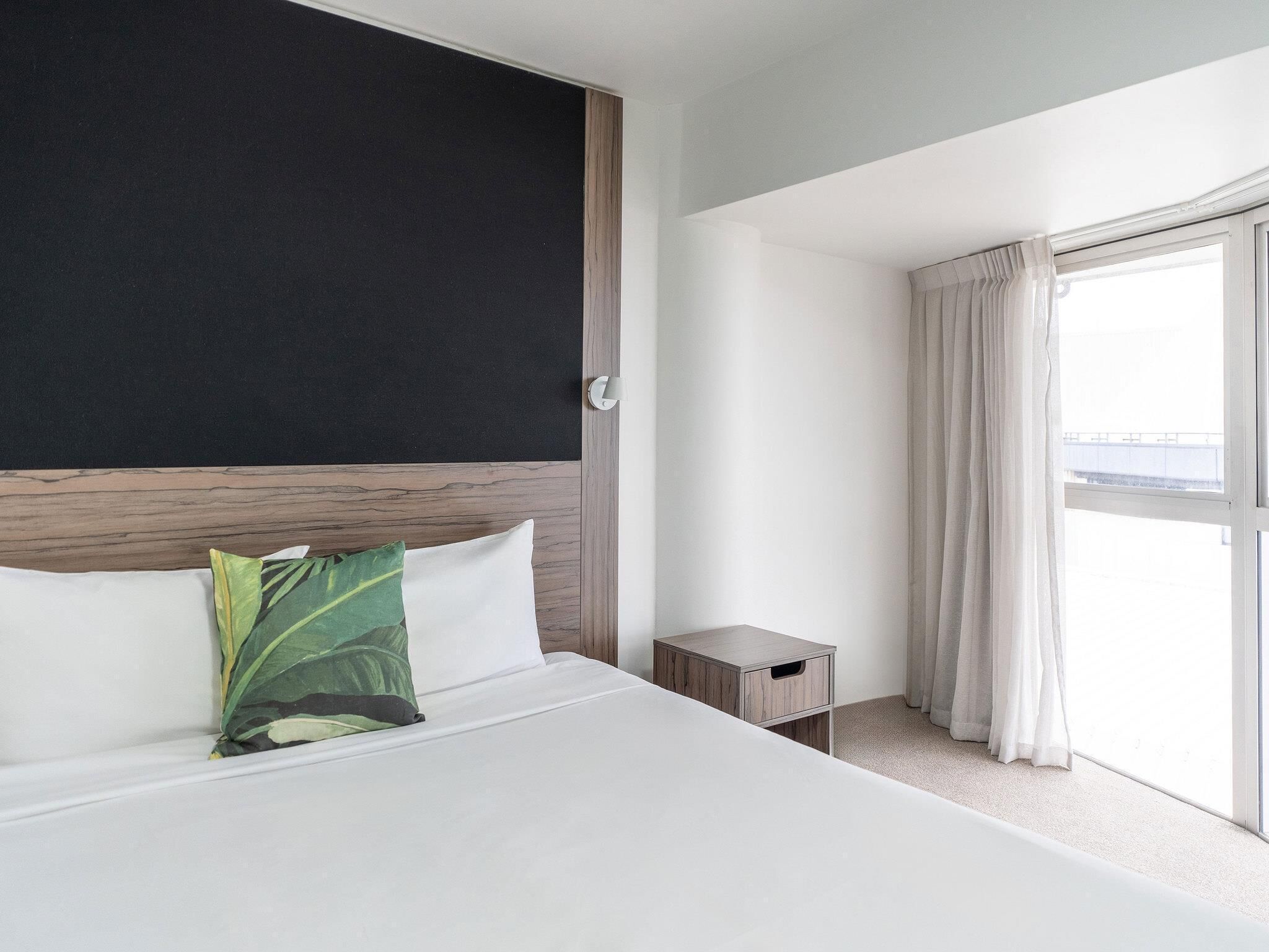 Ein Hotelzimmer mit Bett, Nachttisch, einem Fenster mit Vorhängen und Blick nach draußen.