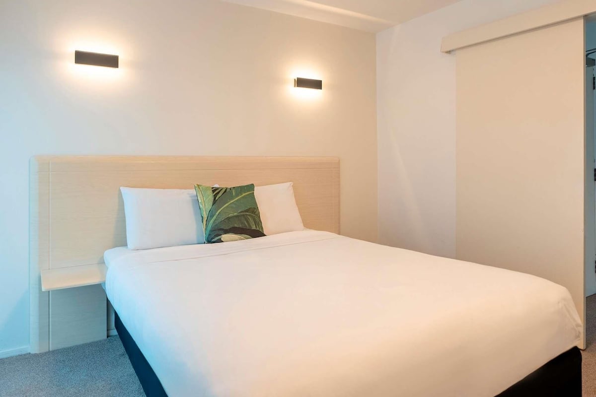Une chambre d’hôtel avec un lit, deux lampes fixées au mur, une table de chevet et une fenêtre avec un store.