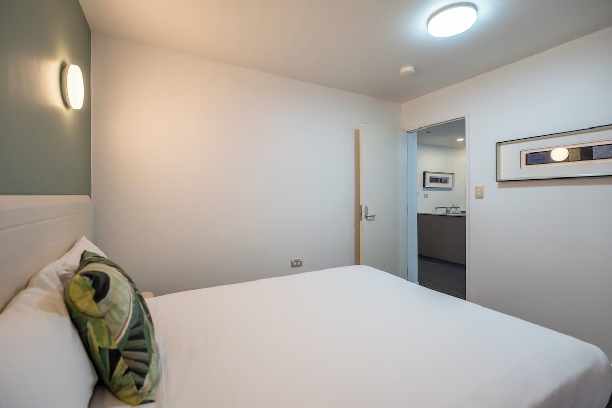 A hotel room with a bed, a pillow, a wall-mounted light, a door leading to a bathroom, and a framed picture on the wall.