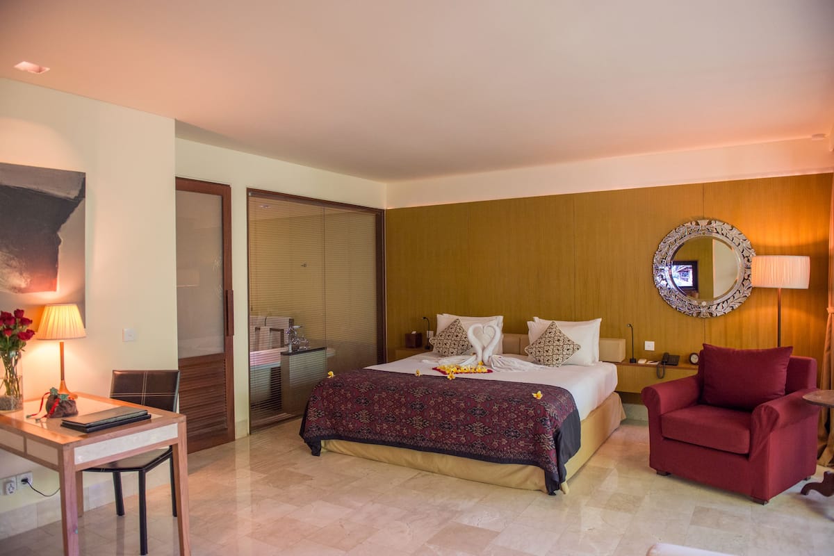 A hotel room with a bed, a desk with a lamp, a chair, a red armchair, and a mirror on the wall.