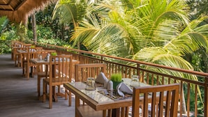 A wooden deck with tables and chairs, surrounded by lush greenery and palm trees. - Komaneka at Rasa Sayang (Ubud)