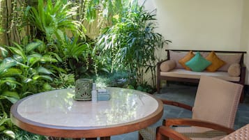 Una mesa redonda de madera con cubierta de vidrio, rodeada de plantas verdes exuberantes. Una silla de mimbre está colocada frente a la mesa.