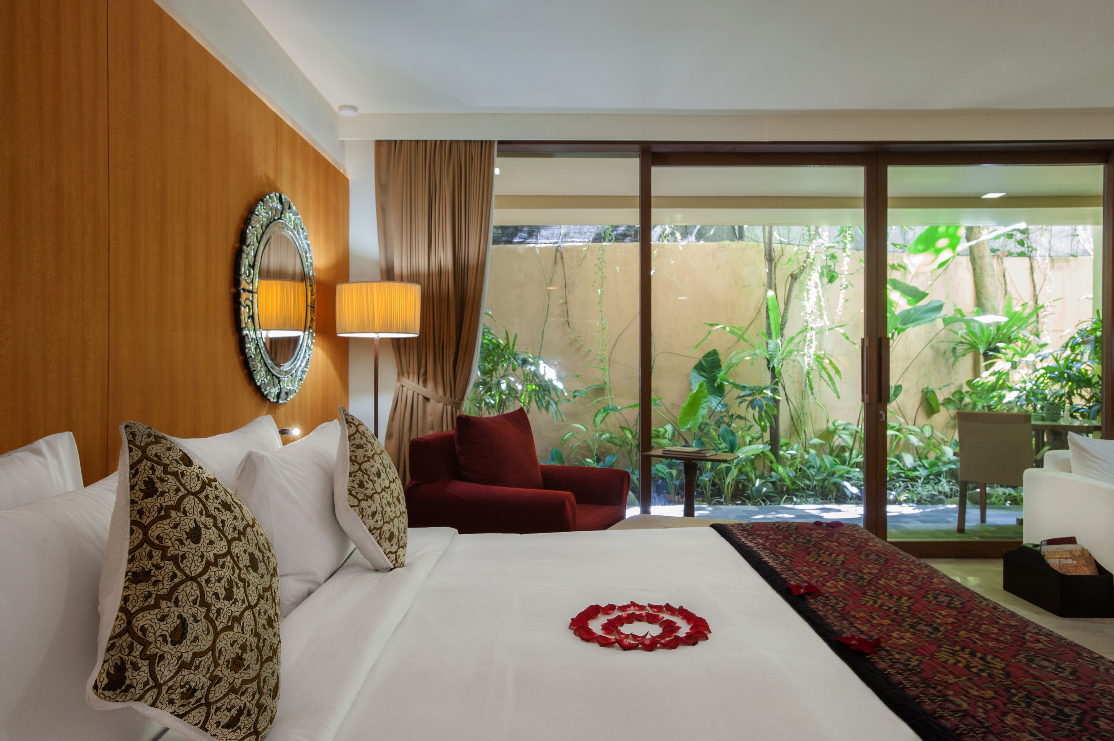 A hotel room with a large bed, a red sofa, a small table, and a view of a garden through a sliding glass door.
