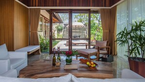 A spacious room with a wooden floor, a large sliding glass door leading to an outdoor area, a sofa, a dining table with a bowl of fruit, and a potted plant. - Komaneka at Rasa Sayang (Ubud)
