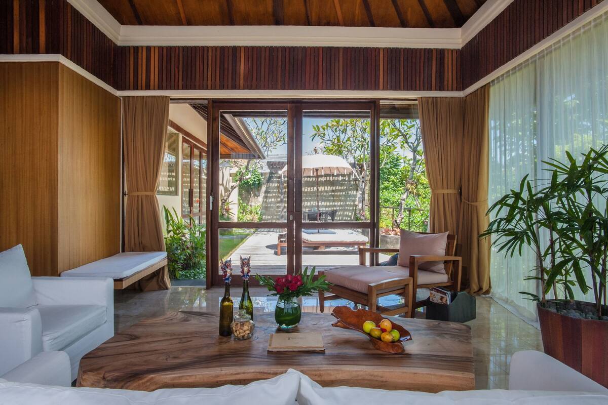 A spacious room with a wooden floor, a large sliding glass door leading to an outdoor area, a sofa, a dining table with a bowl of fruit, and a potted plant.