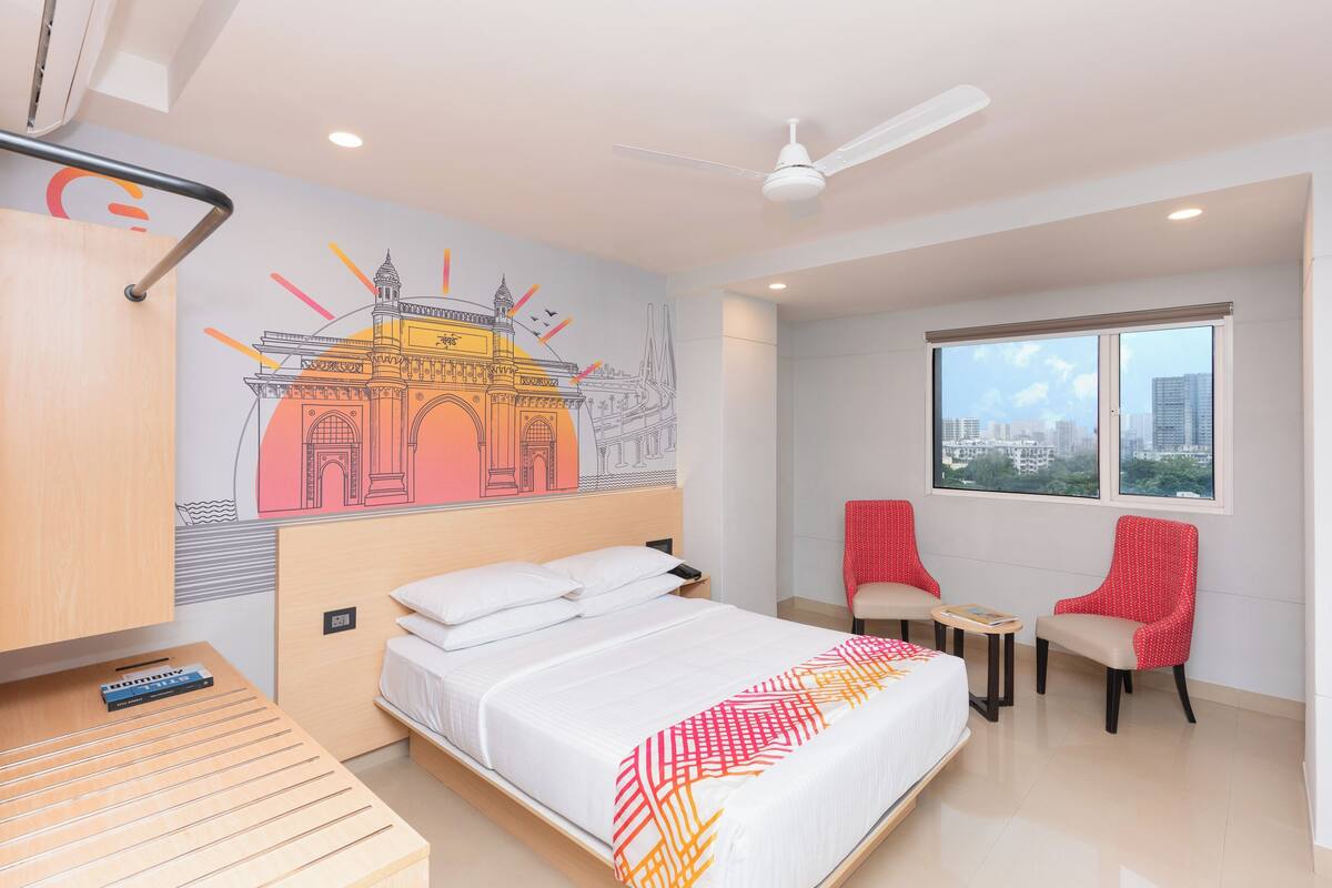 Ein Hotelzimmer mit Bett, zwei Stühlen, einem kleinen Tisch, einem Deckenventilator und einem Fenster mit Blick auf die Stadt.