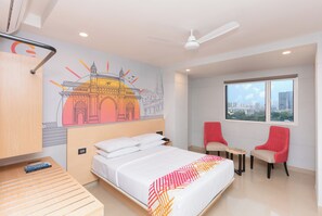 Ett hotellrum med en säng, två stolar, ett litet bord, en takfläkt och ett fönster med utsikt över staden.