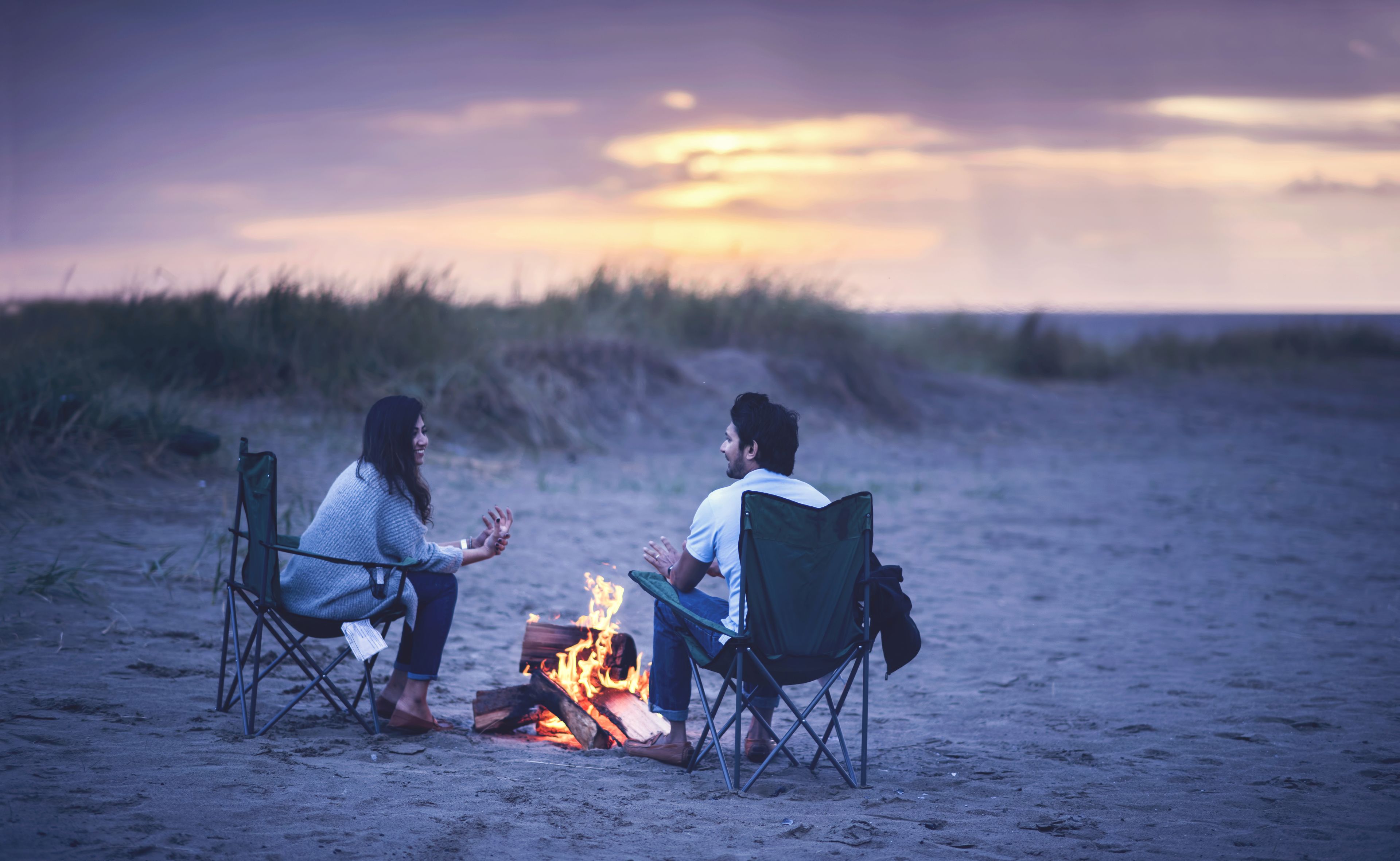 Two people sitting on beach chairs facing a campfire on a sandy beach with a sunset in the background.