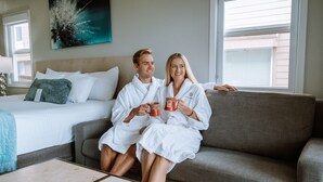 A couple in white bathrobes sitting on a grey sofa in a hotel room with a bed, a window, and a painting on the wall. - Inn at the Prom Oceanfront Seaside (Seaside)