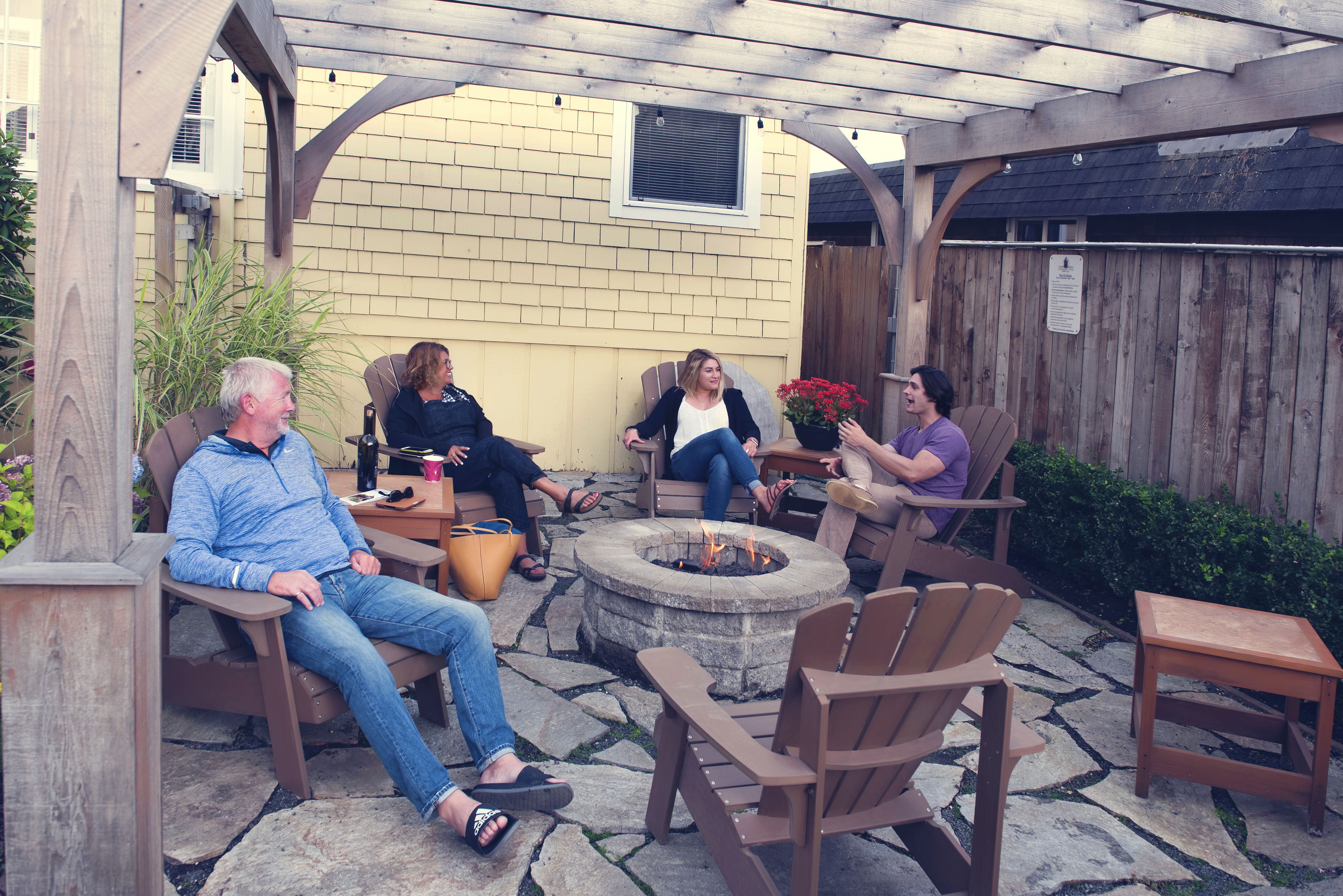 A group of people seated around a stone fire pit in an outdoor patio area with wooden furniture and a pergola overhead.