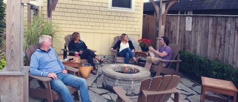 A group of people seated around a stone fire pit in an outdoor patio area with wooden furniture and a pergola overhead.