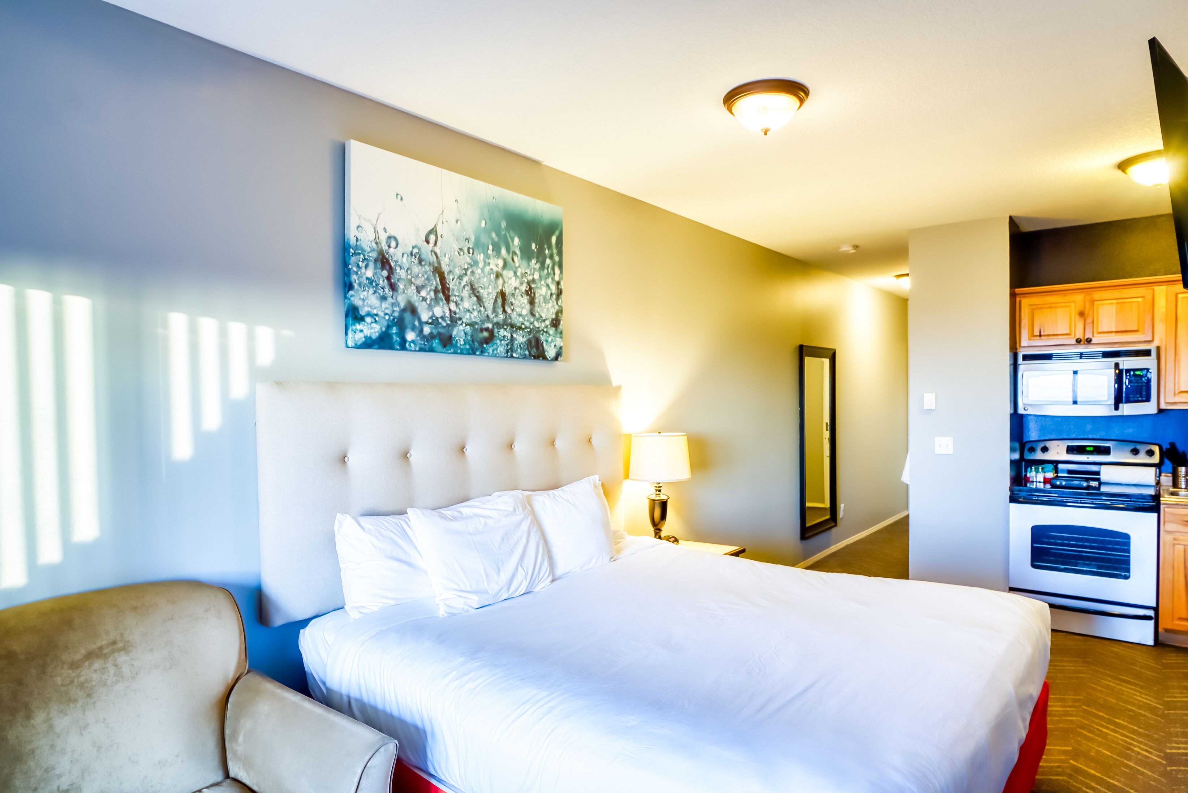 A hotel room with a large bed, a headboard, a painting, a lamp, a mirror, and an adjacent kitchen area.