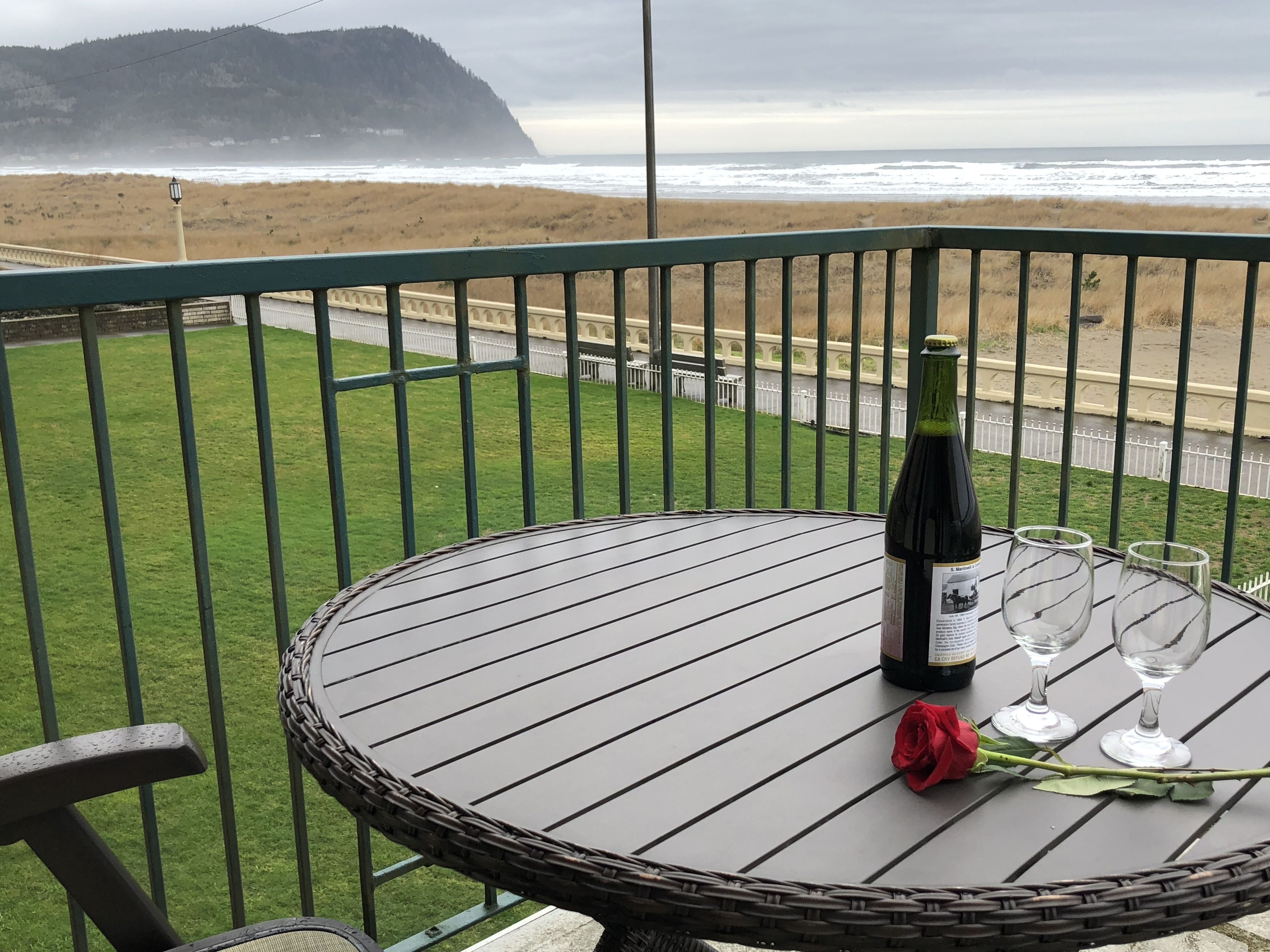 A balcony with a table featuring a bottle of wine, two wine glasses, and a rose. The view includes a beach and ocean.