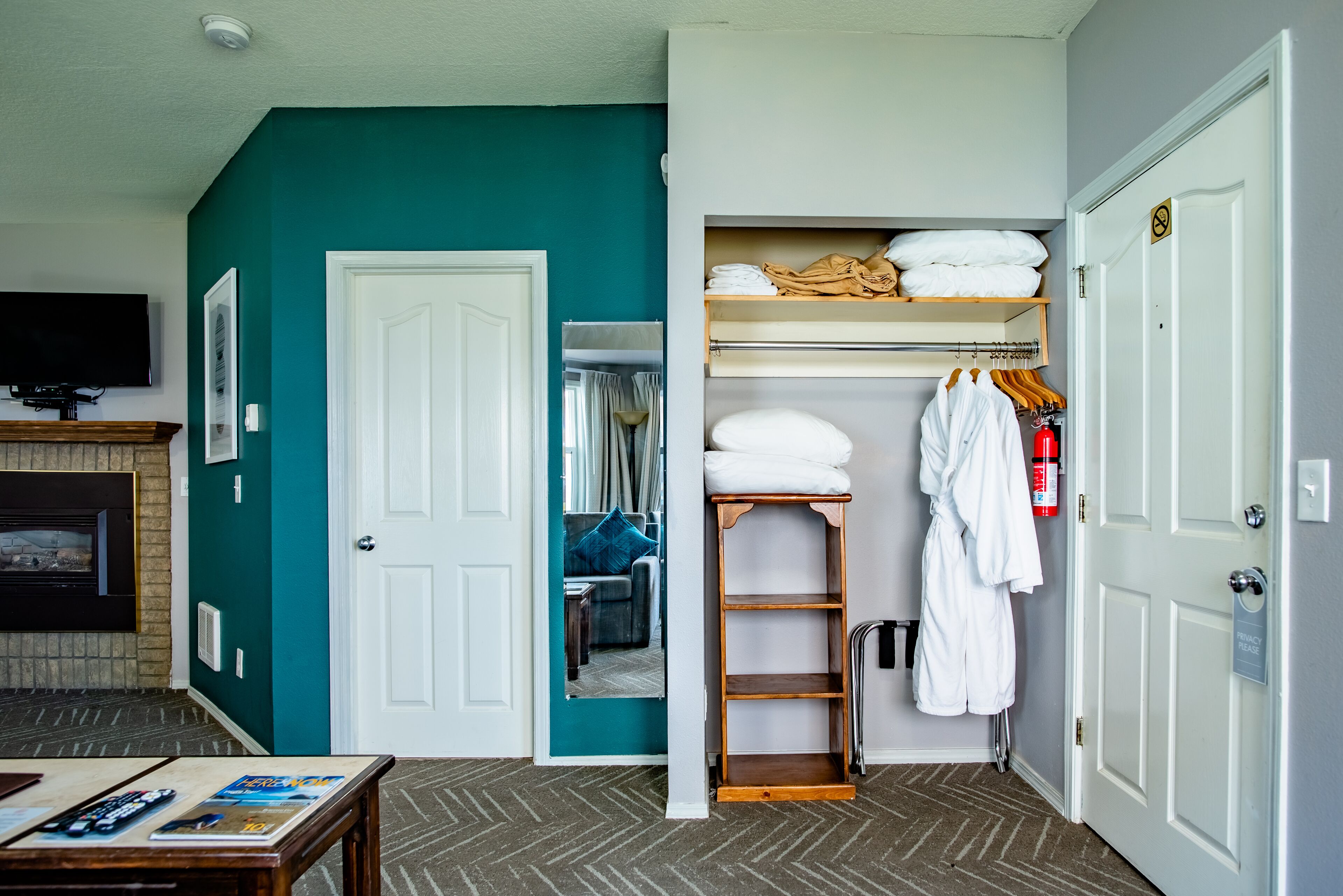 A hotel room with teal walls, a white door, a wooden shelf with towels, a white robe, and a fire extinguisher.