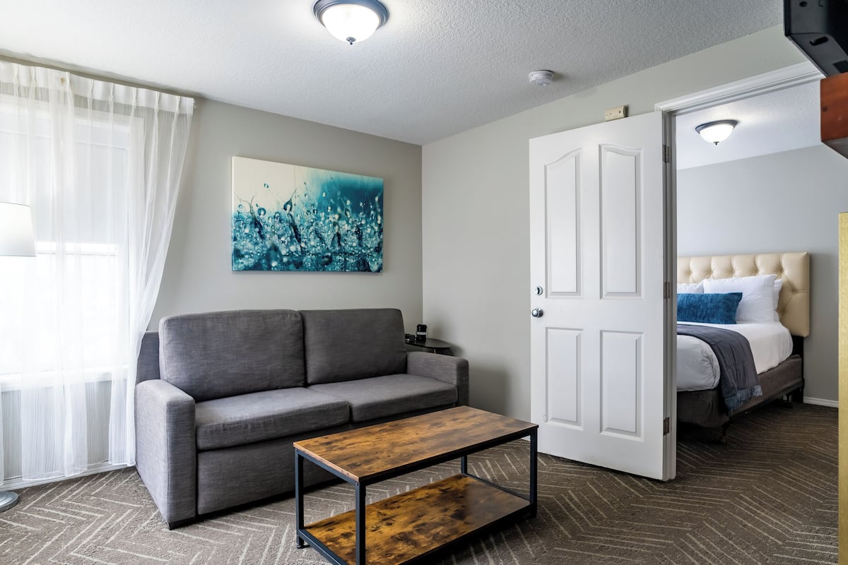 Ein modernes Hotelzimmer mit grauem Sofa, einem Holztisch, einem Bild an der Wand und einem Bett mit einer gesteppten Kopfteil.
