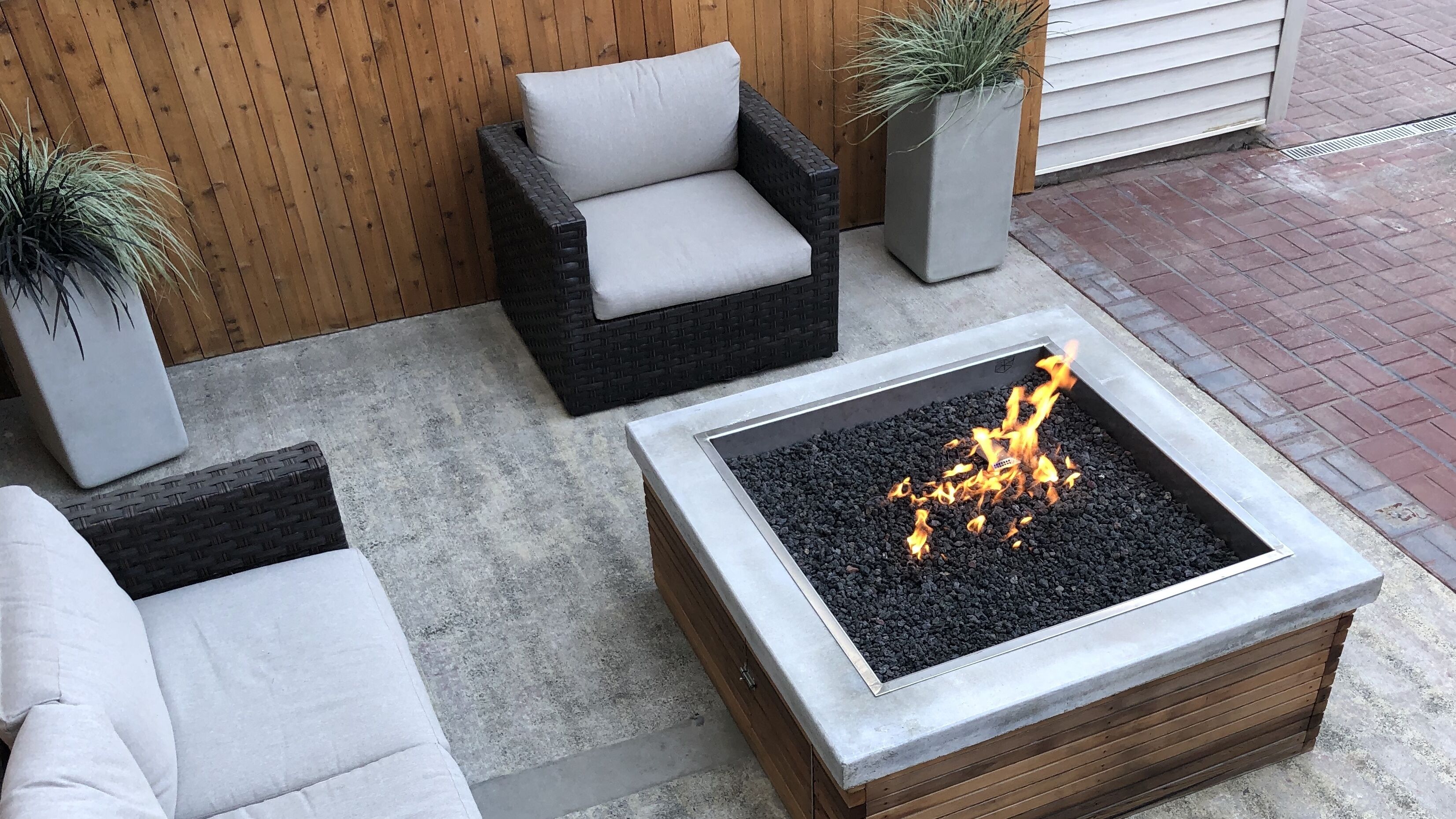 Un espace patio comprenant un foyer, deux fauteuils en osier avec des coussins et deux plantes en pot.