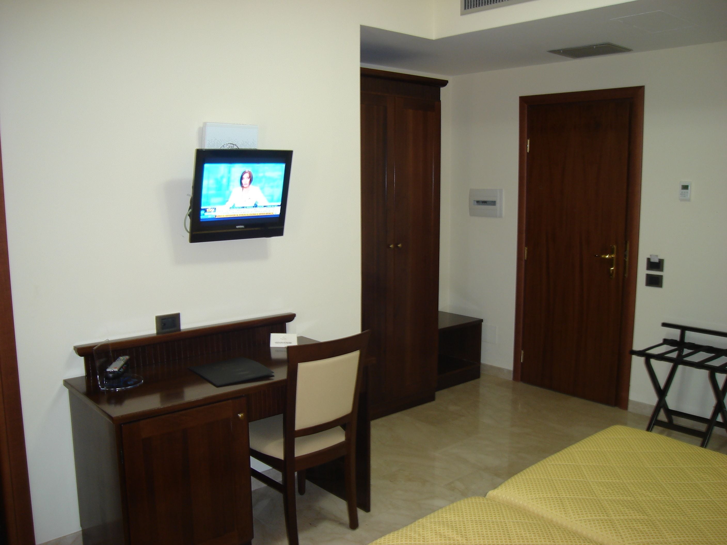 Una camera d'albergo con una scrivania in legno, una sedia, un televisore fissato al muro e un letto con copripiumino giallo.
