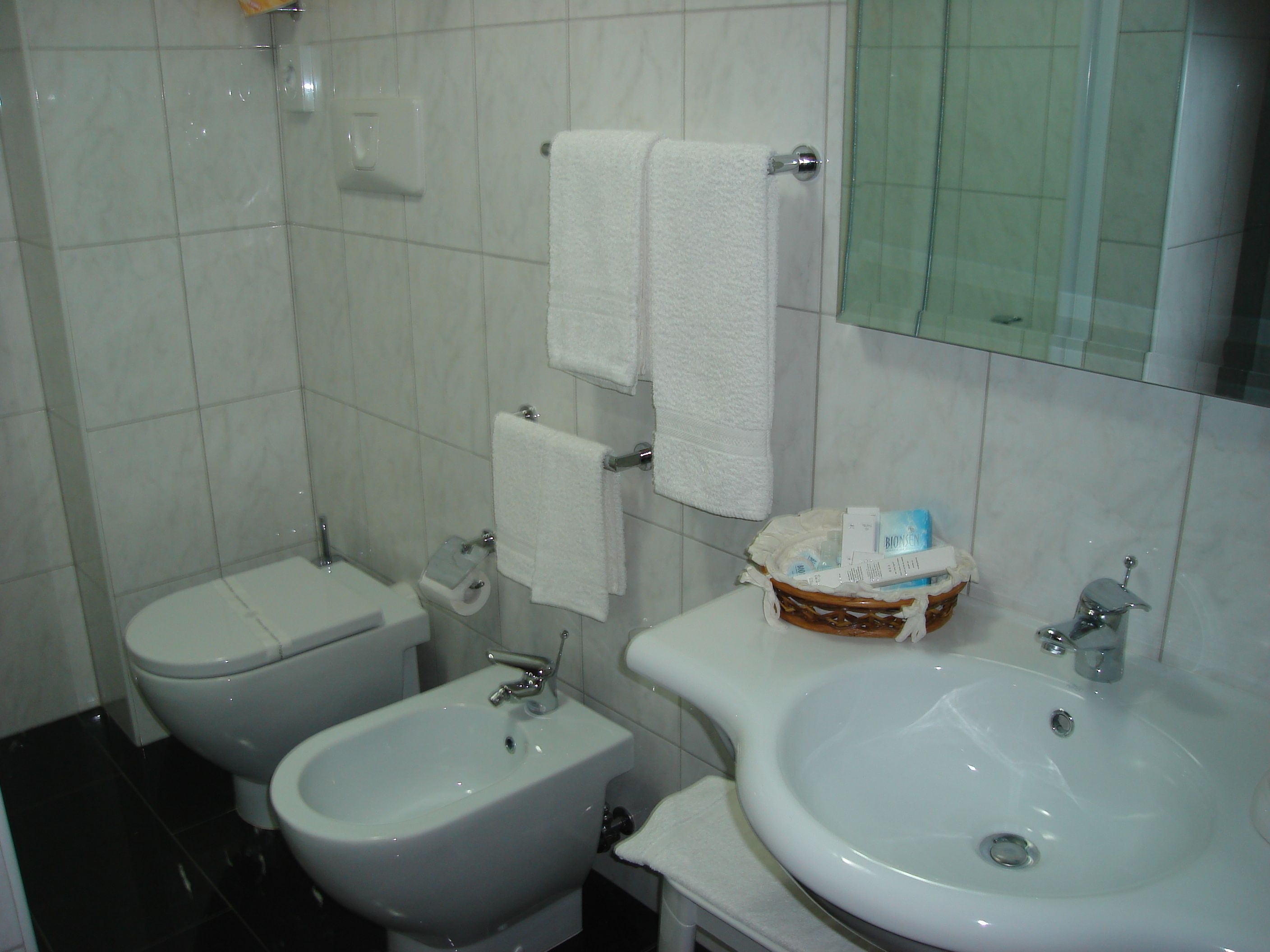 A bathroom with a toilet, bidet, and sink. There are two white towels hanging on the wall.