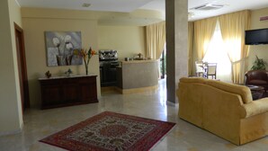 A spacious living area with a yellow sofa, a red patterned rug, a kitchen in the background, and a flat-screen TV mounted on the wall. - Hotel Le Palme (Gioia Tauro)