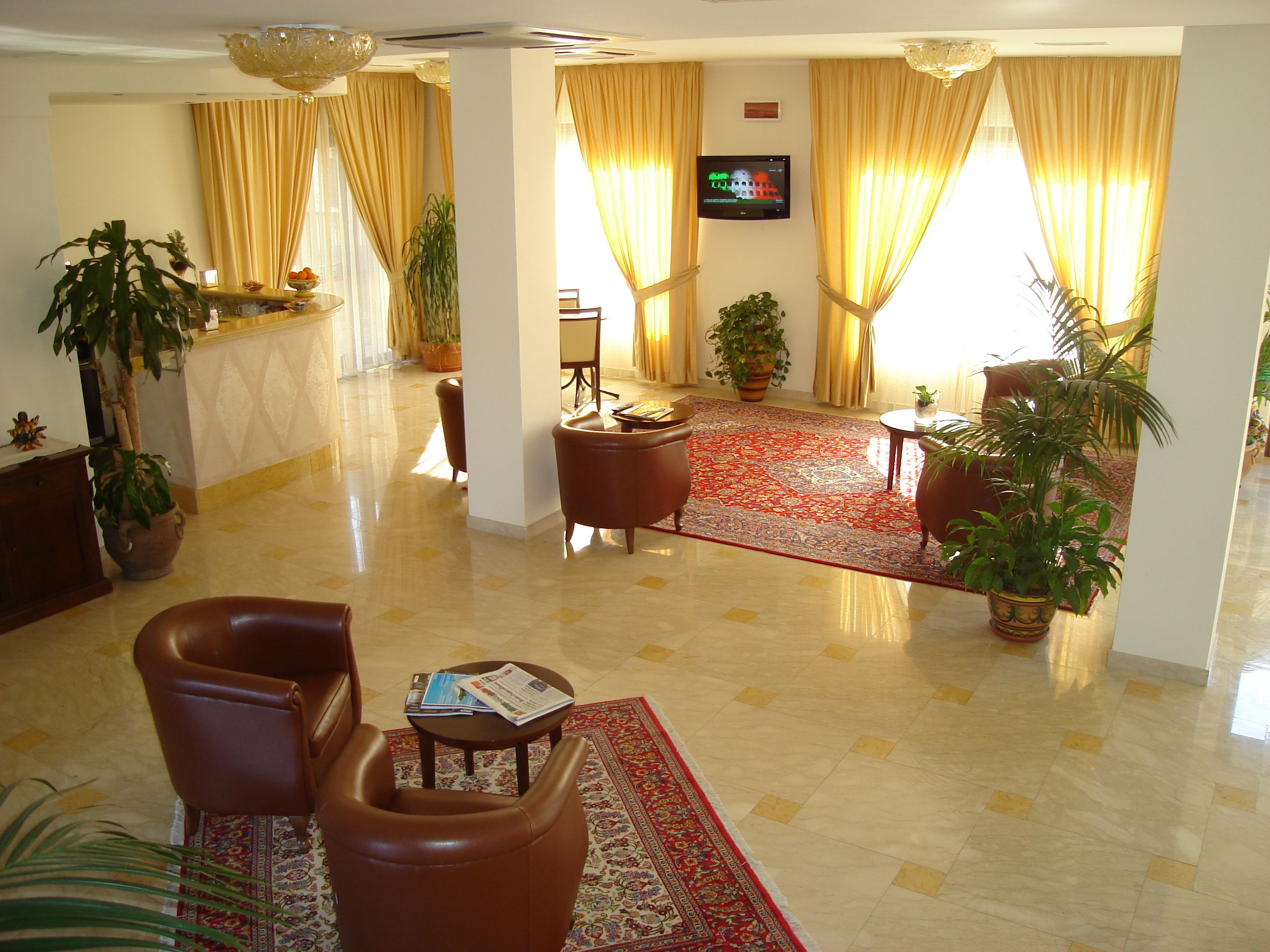 A spacious lobby with a red patterned rug, leather armchairs, a television mounted on the wall, and potted plants.